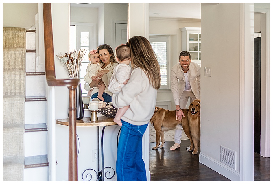 mother holding baby girl in front of a mirror with father and dogs watching
