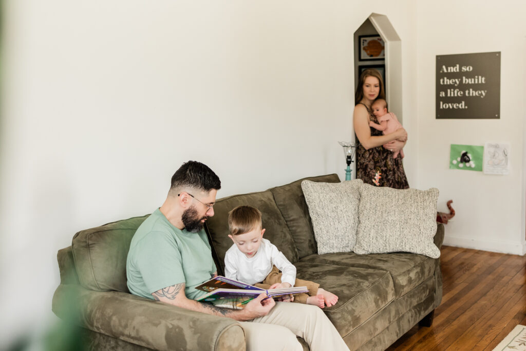 toddler sitting on sofa with his dad looking at a book while mother holds baby and watches lovingly

