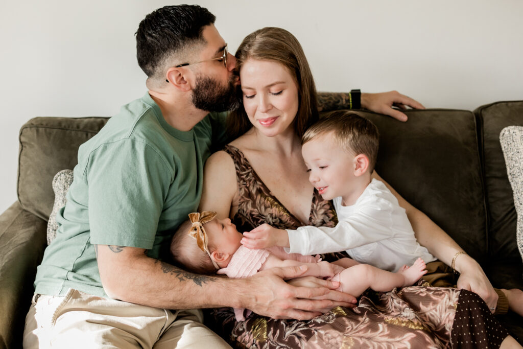  Including Toddlers in Newborn Photo Sessions -toddler looking sweetly at baby sister while sitting on woman's lap on the sofa
