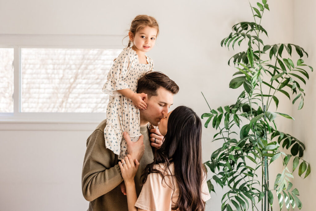 Mom and dad kissing while toddler sits on dad's shoulders